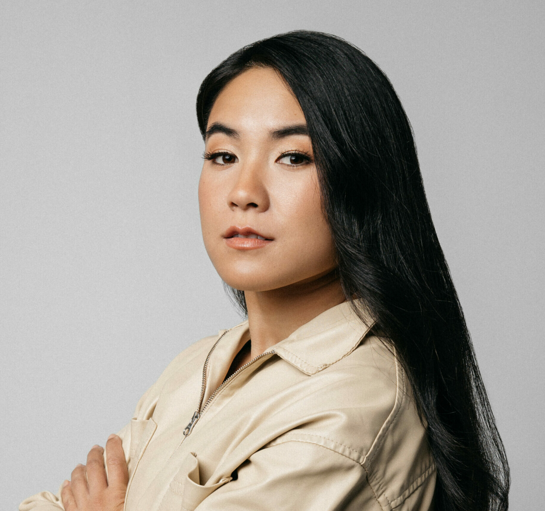 Michaela Ternasky-Holland is a mixed race person with light-tan skin and long black hair that is slightly curled at the ends. She gazes into the camera in a confident and powerful manner with her arms crossed. She is wearing a cream top that zips. The background is a neutral light grey. 