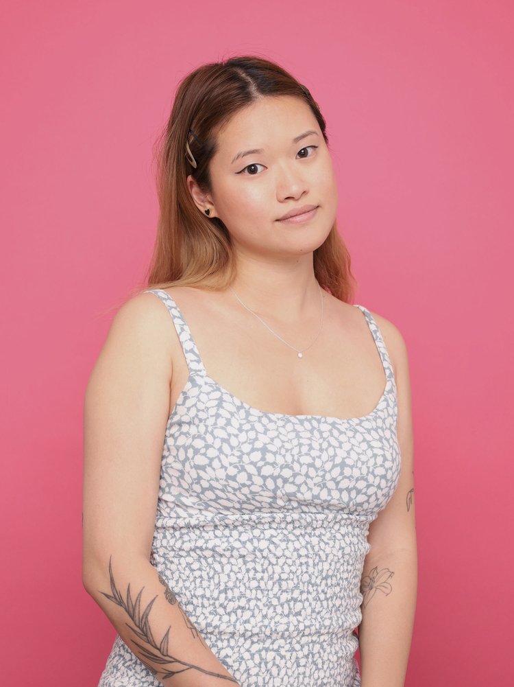 Amanda Lin, a Taiwanese person with light-tan skin and dyed brown hair smiles with her mouth closed at the camera. She is wearing a white and teal sleeveless dress and has black ink tattoos on her arms. The background is pink and her straight hair is tucked behind her ears.