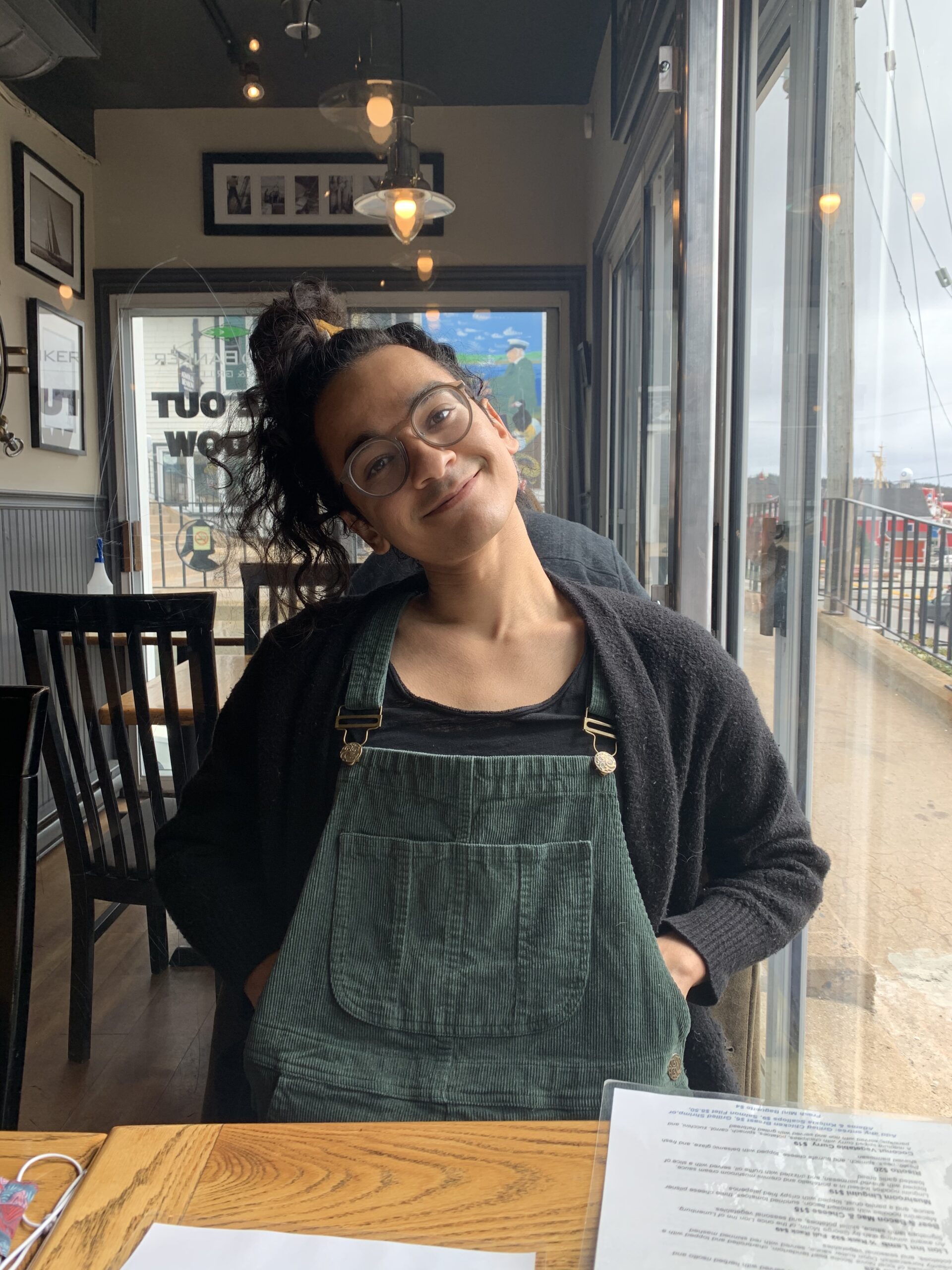 Jake Runeckles, a mixed race South Asian person with round glasses and a top bun is smiling with her head tilted to one side. She is sitting down in a café, wearing green corduroy overalls and a black cardigan, with her hands tucked in her pockets.