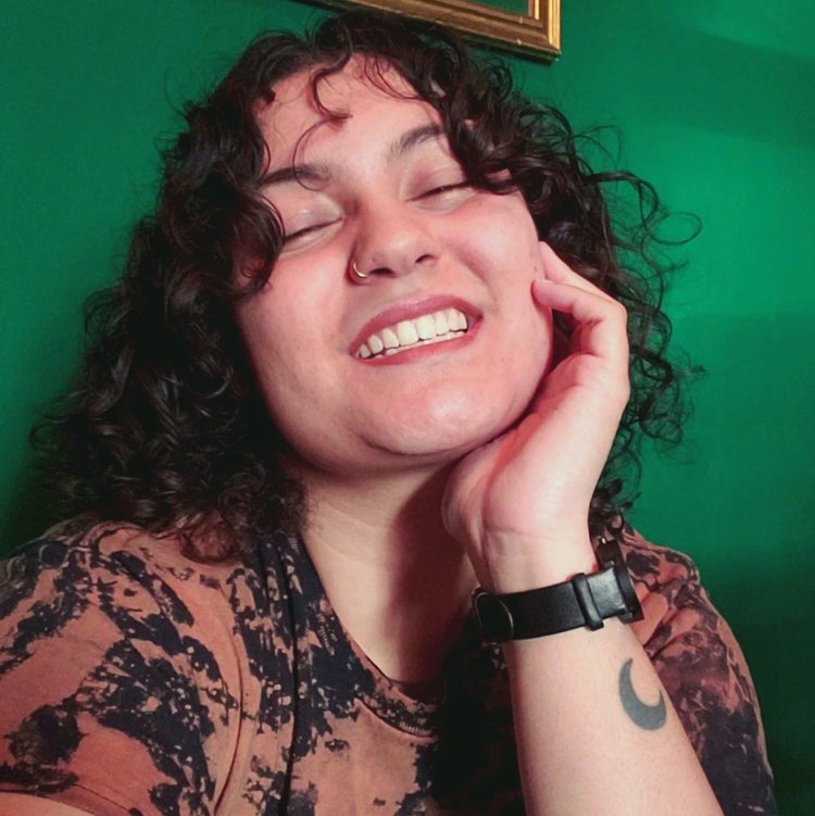 Jasmine Noseworthy Persaud, a mixed race Guyanese person with light brown skin and dark curly hair smiles wide with teeth showing and hand holding their face. They are in front of an emerald green wall and wear a brown-black tie-dye shirt.