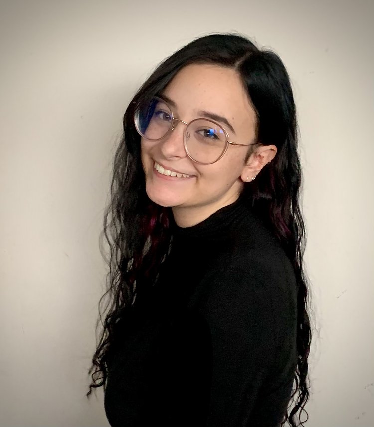 Justine Katerenchuk, a white woman with deep brown hair smiles with teeth as she's looking into the camera. She has large round metal thin framed glasses with long deep brown wavy hair. She’s standing in front of a white wall.