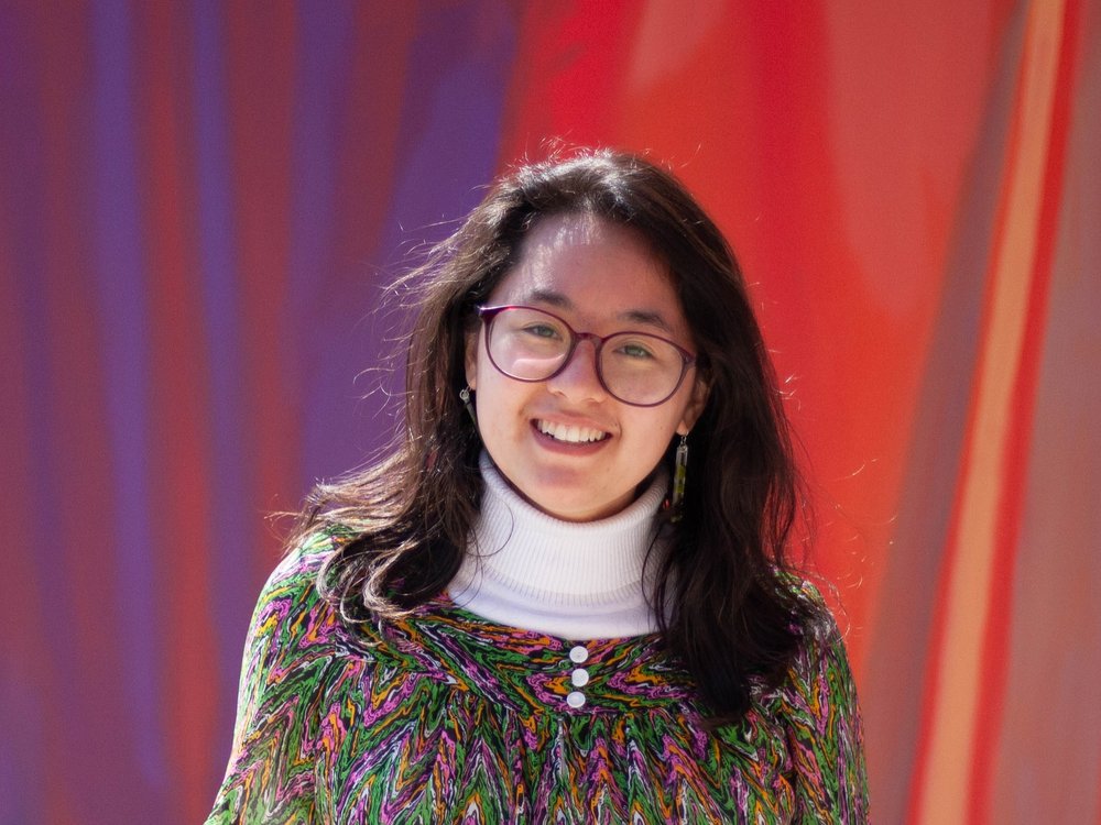 Nicole Eun-Ju Bell, a mixed race Korean-European woman with dark brown hair, smiles at the camera. She has red frame glasses, and is wearing a white turtleneck under a brightly colored shirt that has zigzags of green, pink, and orange. The background is a red and purple abstract mural.