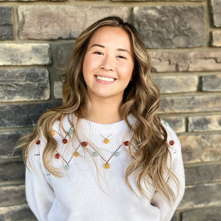 Samantha Vu, a Chinese woman with curly dark brown and blonde hair is smiling at the camera. She is wearing a white sweater with red yellow and blue pom poms and her arms are down. She is standing in front of a brick wall.