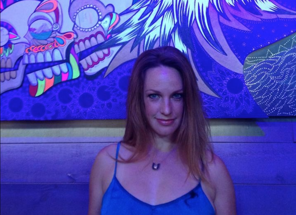 Lee Erdman, a light skinned Italian-Danish Canadian person with long, strawberry blonde hair, smiles with her mouth closed at the camera. Her background is indigo blue and purple.