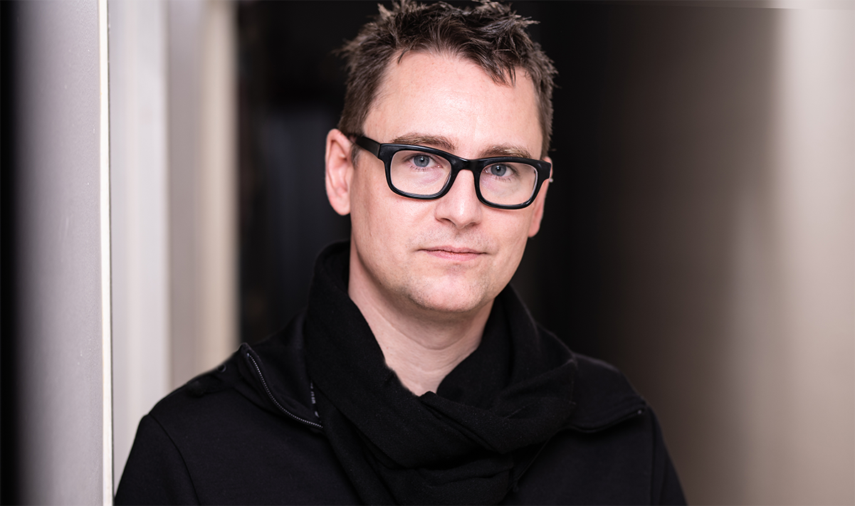 Professional headshot of a nonbinary white person looking at the camera. They have blue eyes, short-cropped brown hair and wear thick-rimmed black glasses. They are wearing black and standing against a dark, abstract background.