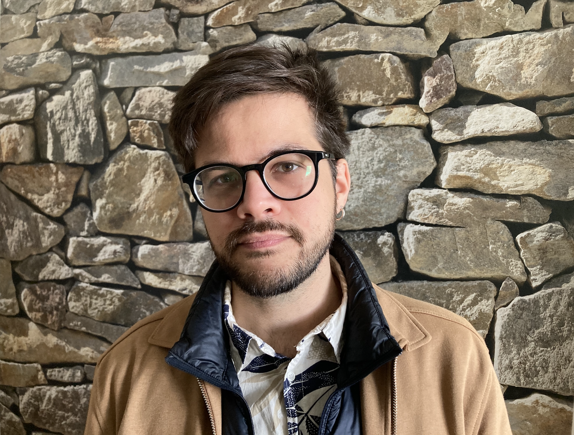 Profile shot of a Latino man with short dark brown hair and beard. He wears corrective glasses with black frames and a pair of jackets (dark blue and light brown) layered over a white shirt with leaf patterns. He looks at the camera half smiling. He is standing in front of a rock wall.
