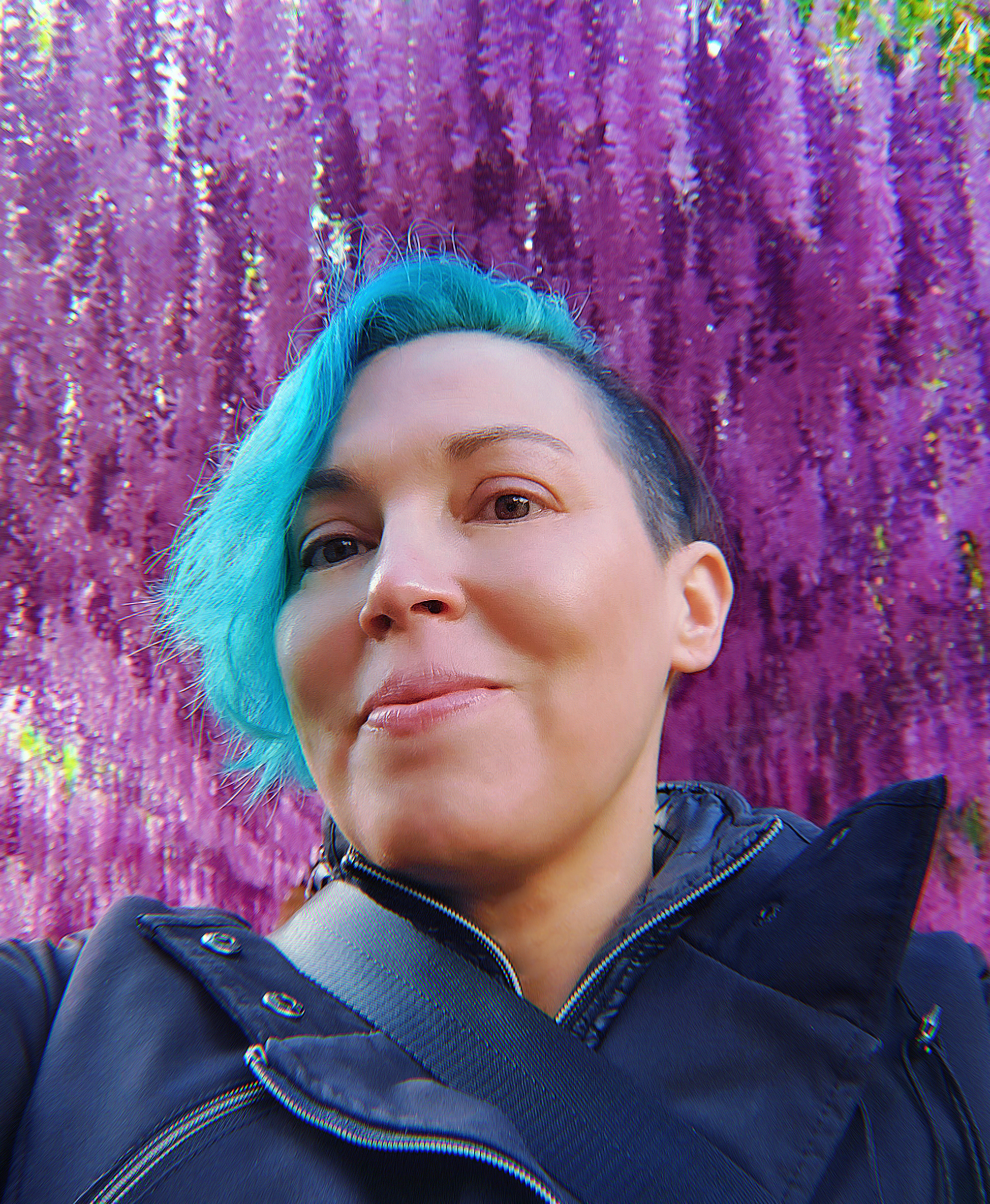 A Native person with light skin and blue hair with shaved sides, smiling. They are wearing a black rain jacket and standing under a tunnel of vibrant purple wisteria flowers.