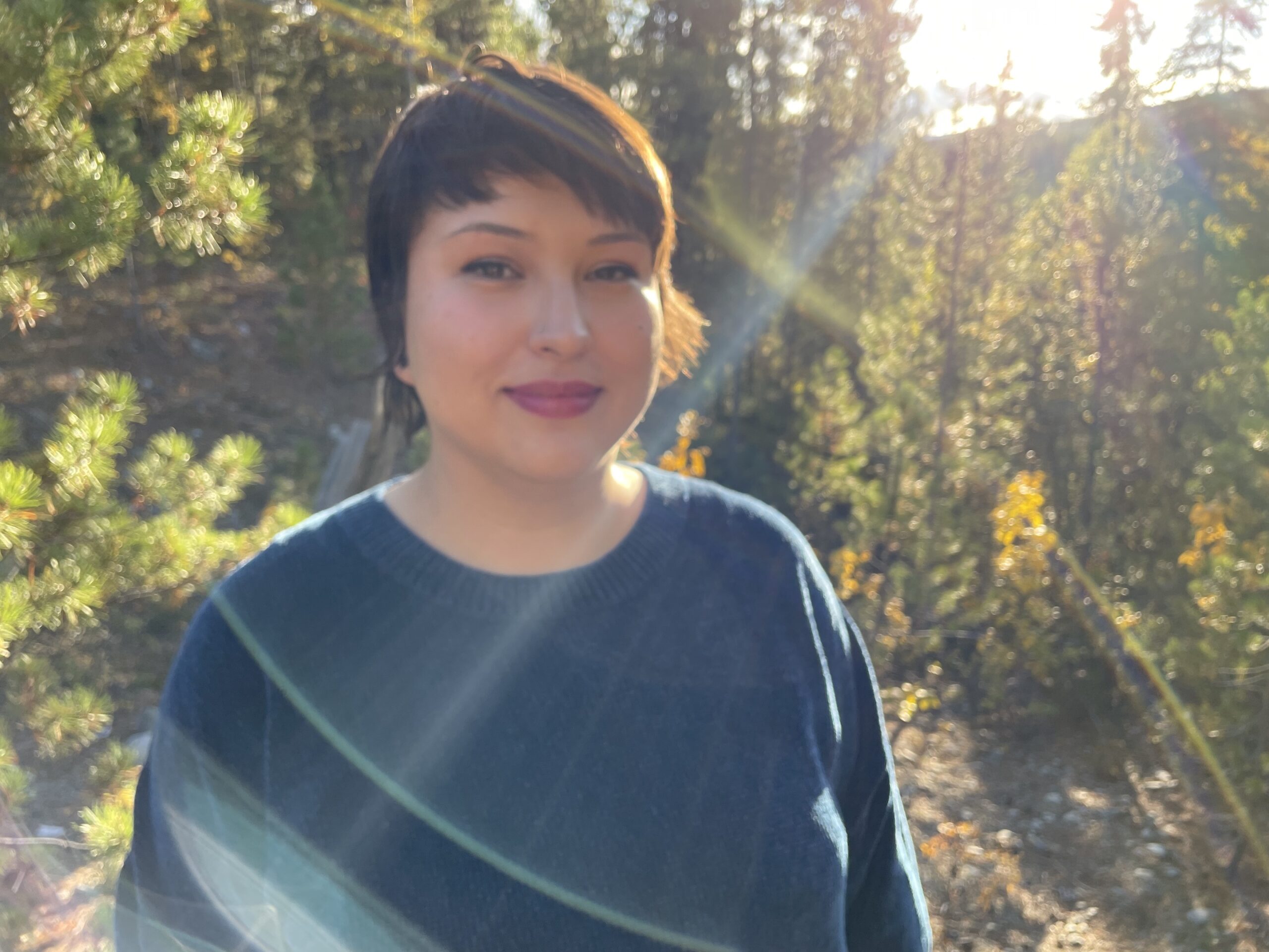 Heather ‘Von’ Steinhagen, a Cree/German person with light skin, brown hair, and dark eyes with her mouth closed, smiling at the camera. She is wearing a fuzzy blue sweater outdoors in a sunny boreal forest. There is a blue sun glare near her face.