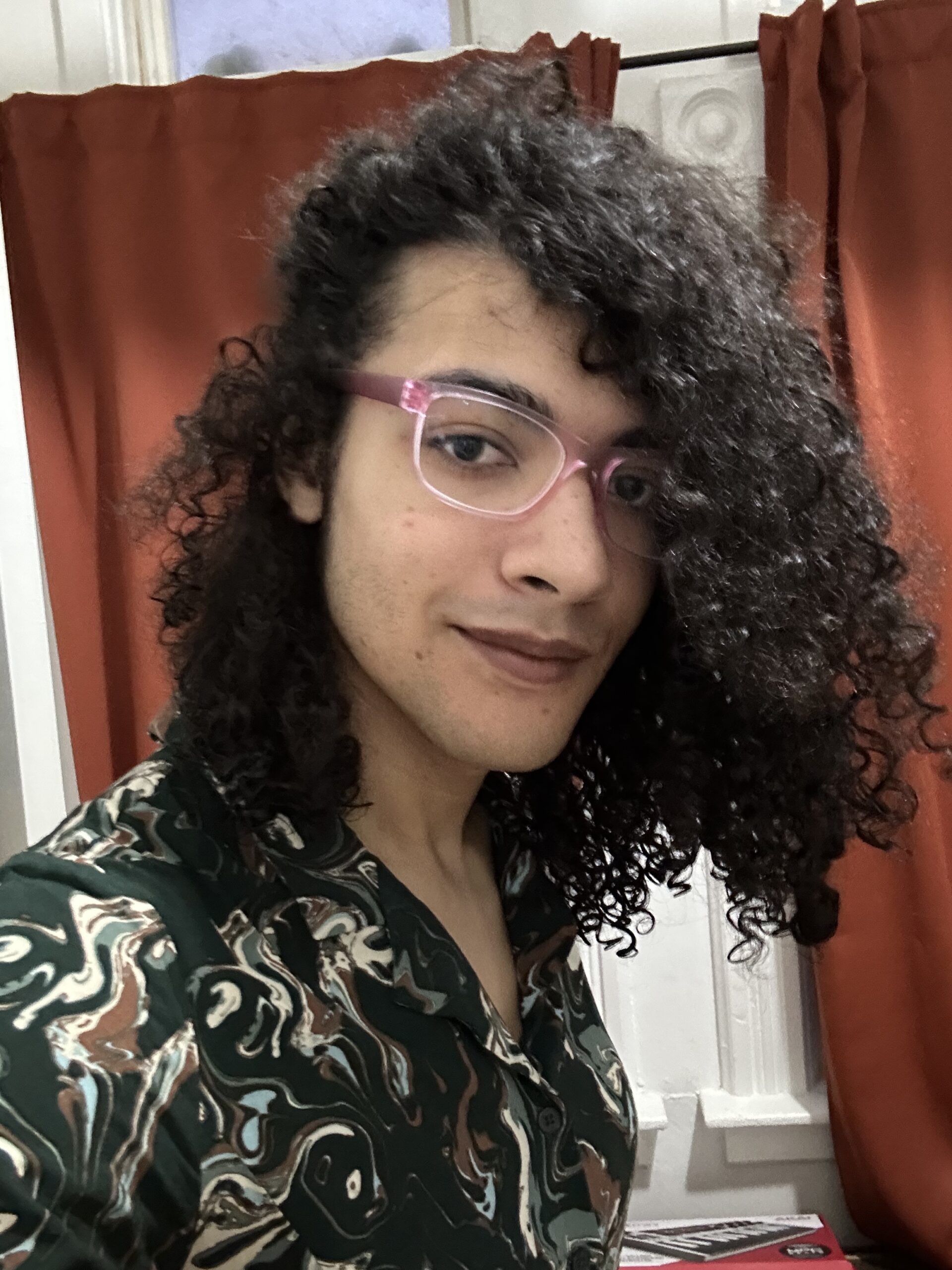 A light brown-skinned trans woman wearing a leafy-patterned dark green dress, pink square glasses, and brown lipstick leaning to the camera in 3/4 view with a slight smile. She has shoulder length, fro'ed black hair.