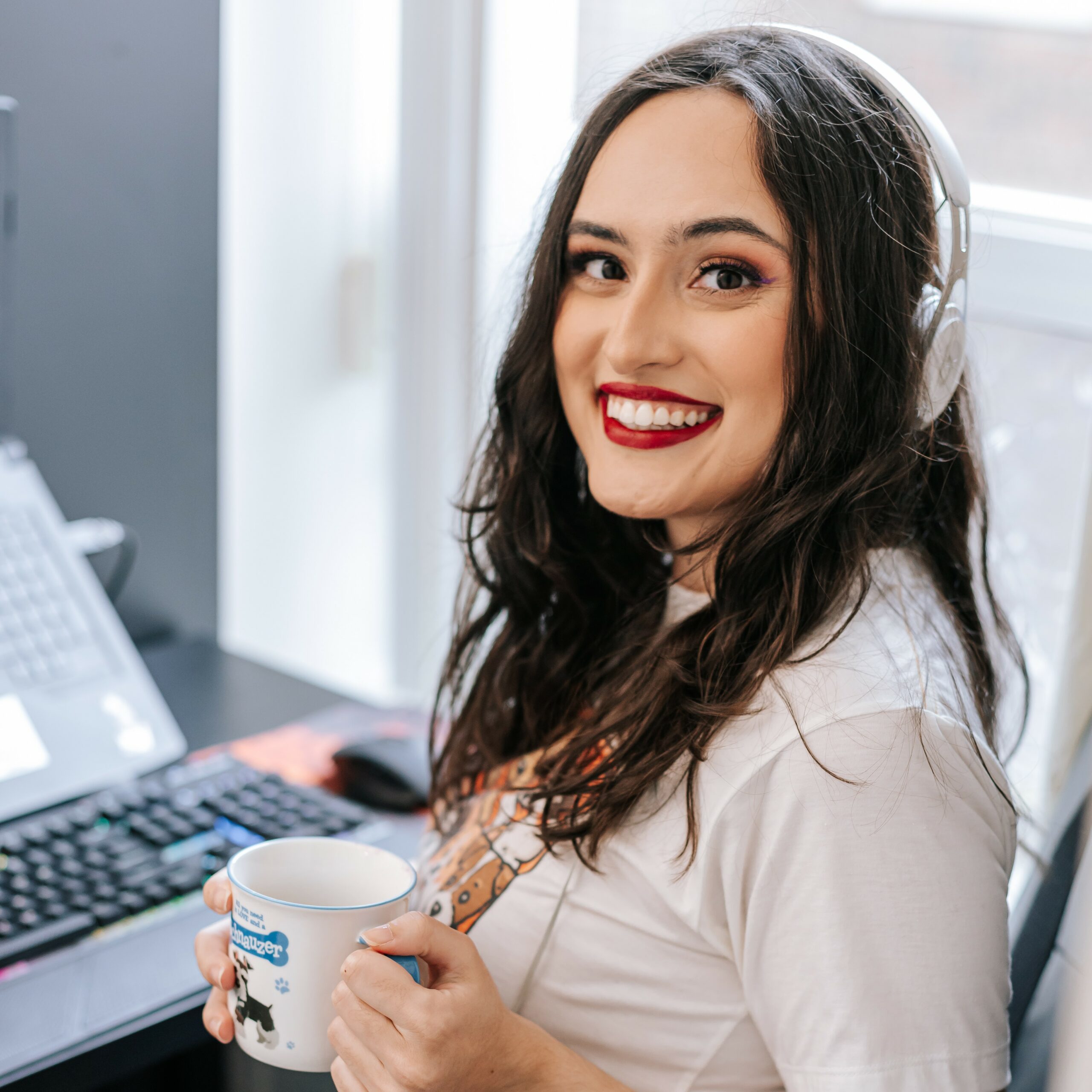Amanda Duarte, a woman with light skin and dark brown hair smiles with her mouth open at the camera. She is wearing a white t-shirt with dog drawings on the front and holding a white and blue mug with both hands. The background has a white window on her right and a black desk with a laptop and keyboard on her right, and she has white headphones on her head.