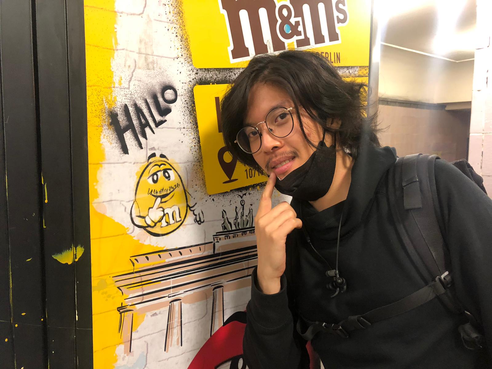 A black-haired Chinese man wearing all black, with round glasses and a thin moustache, holding his finger up his index finger beside his mouth. He is standing in front of a vending machine with a m&m logo, with a yellow m&m character who has been graffiti'd to also have a thin moustache, and who also has his index finger held up beside his mouth. The word HALLO is written above the character.