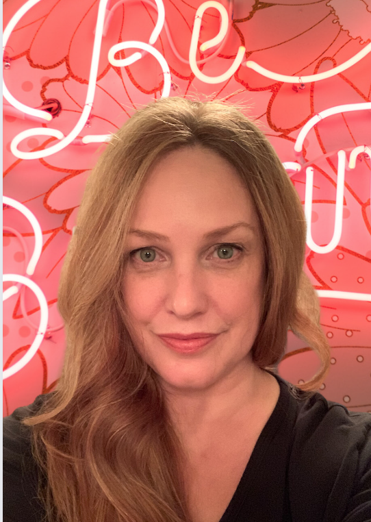 Lee Erdman is an Italian/Estonian Canadian with light skin and strawberry blonde, shoulder length hair. She is smiling in a friendly way in front of a pink backdrop with a neon sign that reads 