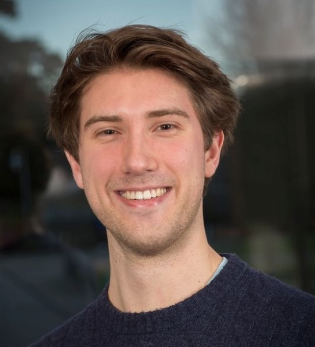 The image shows a young Caucasian man with light brown hair and a friendly smile. He is wearing a casual dark blue sweater and the photo is taken outdoors with a blurred background that suggests a suburban setting. His expression is open and approachable.