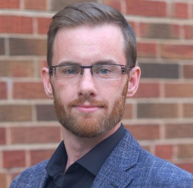 A 30-year white man wearing glasses, with short brown hair and a reddish-brown beard is in front of a red brick wall. He's looking directly at the camera with a slight smile, wearing a light blue textured suit jacket with a dark blue shirt underneath.