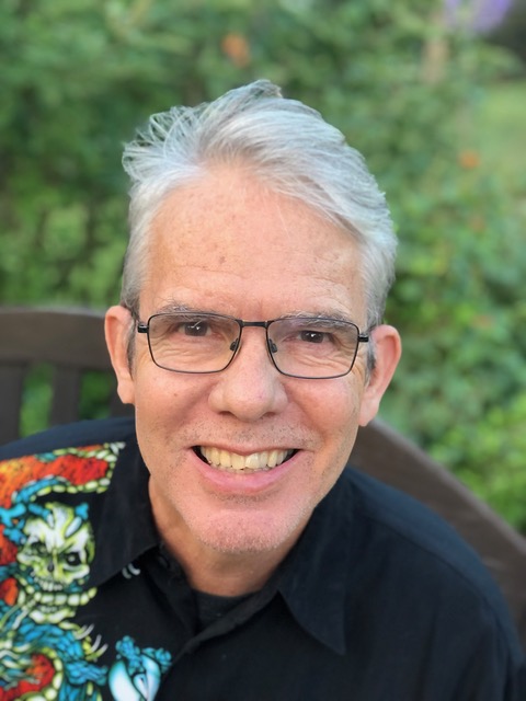 The image shows an older Caucasian man with white hair and glasses, smiling warmly. He is wearing a black shirt with a colorful graphic design, and is seated outdoors with a green, leafy background. His expression is friendly and engaging.