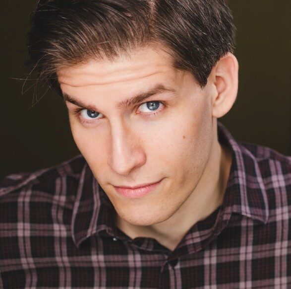 The image features a young Caucasian man with dark brown hair styled neatly to the side, showcasing intense blue eyes. He is wearing a dark plaid shirt and posing against a muted, dark background. His expression is thoughtful and slightly intense, with a direct gaze that engages the viewer.