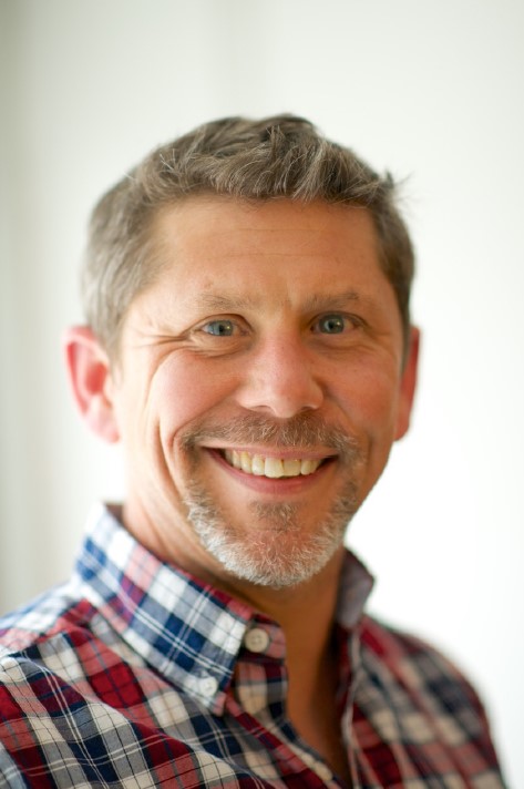 The image shows a middle-aged Caucasian man with graying hair, smiling warmly. He is wearing a plaid shirt with a red and blue pattern. The photo has a soft focus, highlighting his friendly expression in a well-lit setting.