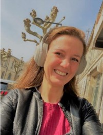 A white woman with brown hair smiles with teeth as she's looking into the camera. She has light grey headphones on and is wearing a black jacket and a red shirt. She is outside in a sunny street, you can see the blue sky and part of a tree behind her.