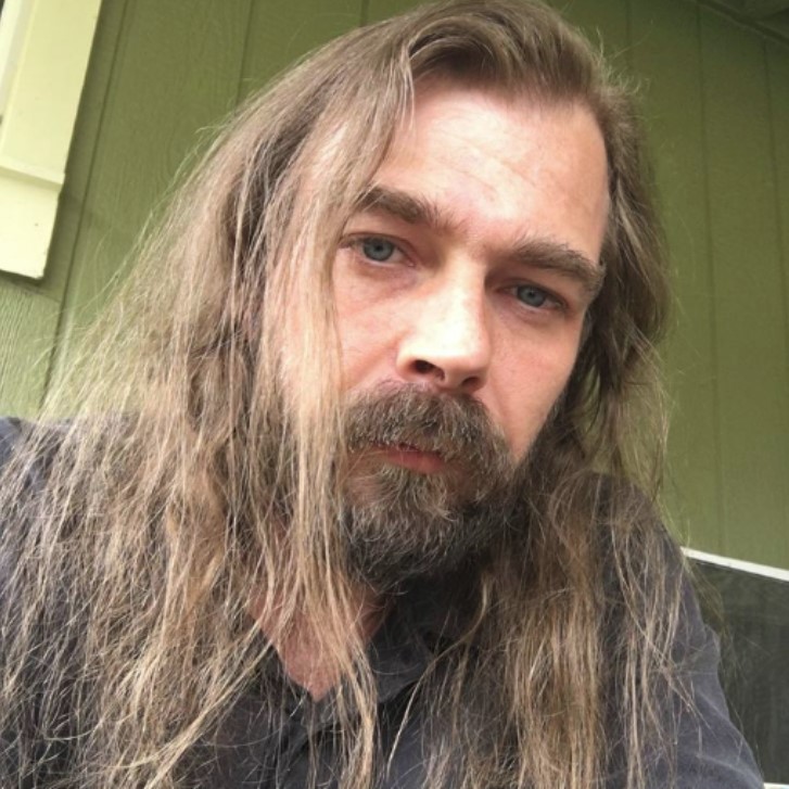 The image shows a middle-aged Caucasian man with long, straight gray hair and a serious expression. He has a full beard and is staring directly at the camera. The background suggests a relaxed, outdoor setting, enhancing the natural and introspective vibe of the portrait.