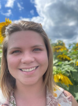 A white early 30s woman with dark blonde hair smiles with teeth and big cheeks and crinkled eyes as she's looking into the camera. It is a sunny day with clouds and sunflowers in the background.