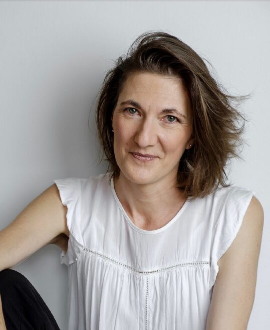 The image features a middle-aged Caucasian woman with shoulder-length brown hair, gently smiling. She is wearing a simple white blouse and is seated in a relaxed posture against a plain white background, giving the portrait a clean and professional look. Her expression conveys confidence and approachability.