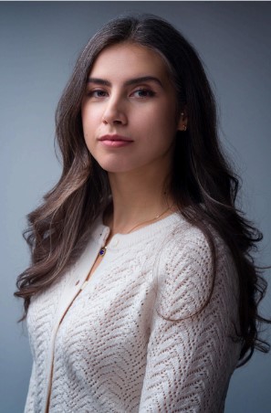 A middle-eastern woman stands with a calm expression. Her long, dark hair falls in soft waves, framing her face. She wears a cream-colored sweater, intricately textured, with cream coloured buttons. The lighting highlights her features, illuminating her clear skin and composed demeanor. Her gaze is steady, reflecting an inner strength. Behind her, a neutral gray backdrop draws attention to her presence.