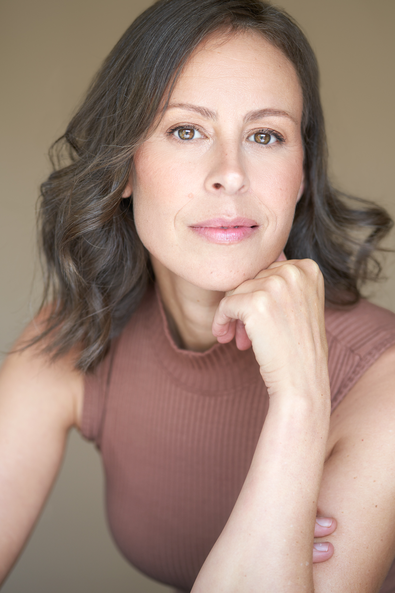 Nita Bowerman, a fair skinned and brown eyed woman with brown wavy shoulder-length hair smiles slightly with her mouth closed. She looks directly at the camera while resting her chin on the back of her curled fingers on her left hand. She is leaning forward slightly. She is wearing a light brown ribbed mock neck tank top. The background is a neutral beige colour.