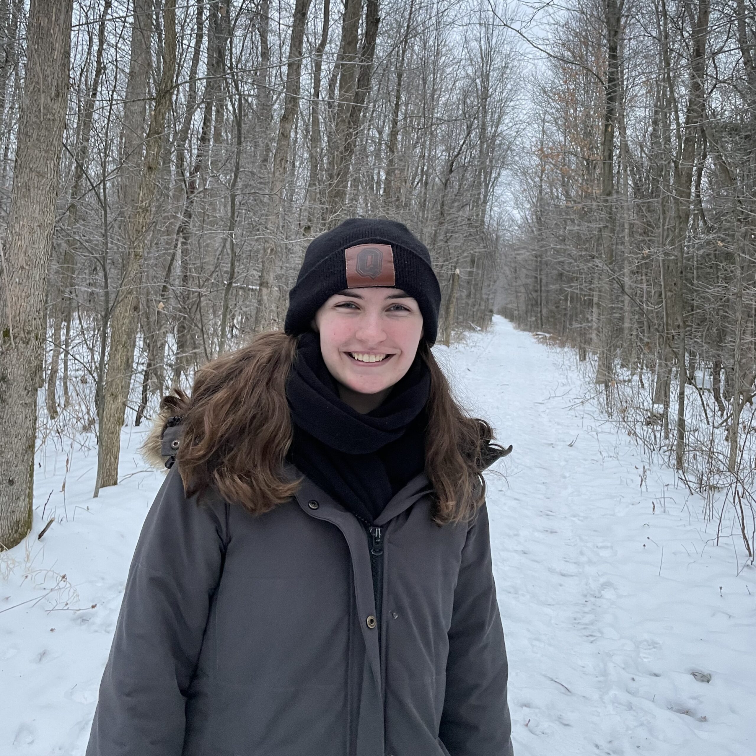 Michaelah Wales, a person with light skin and light brown hair smiles at the camera. She is wearing a grey-brown coat, a black scarf, and a black toque. Her hair covers her shoulders. The background is a forest in wintertime, the ground is covered in snow.