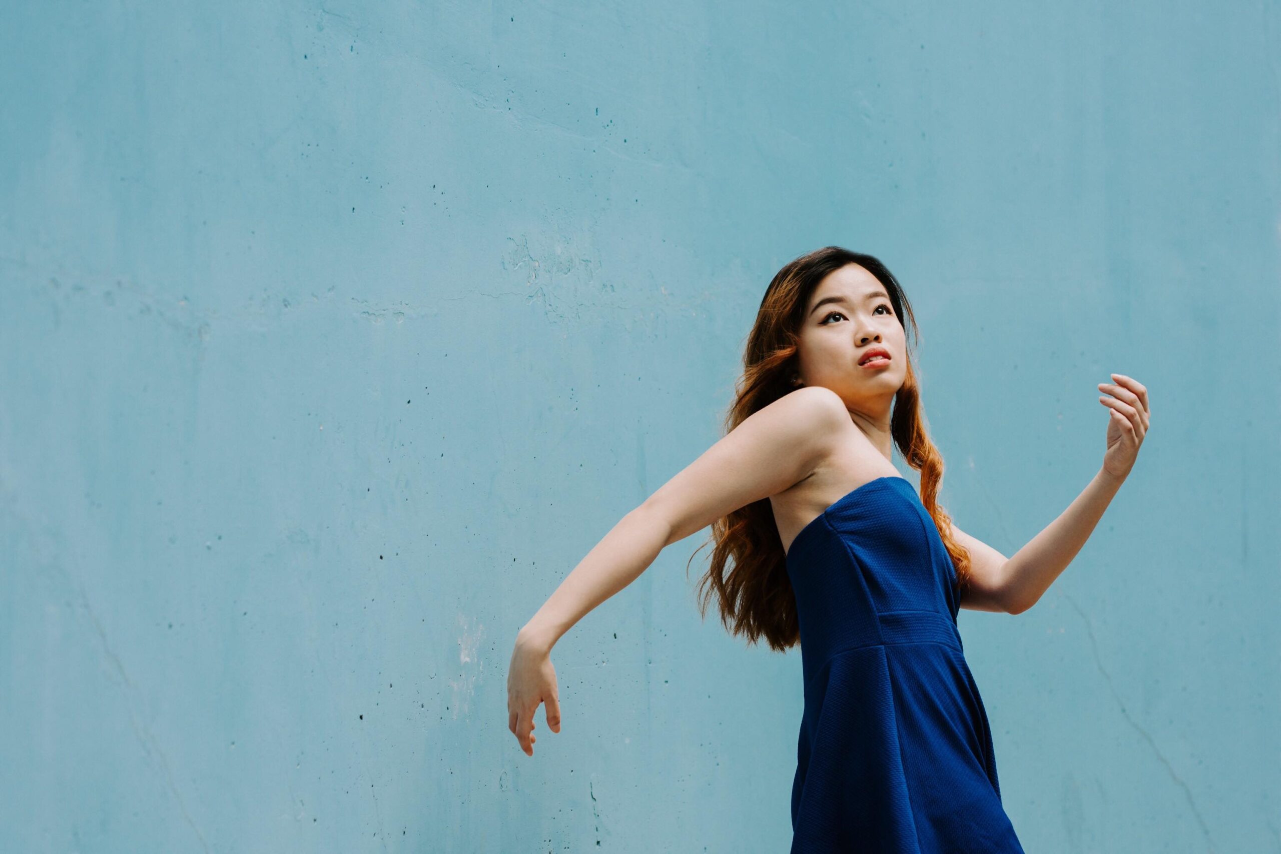 A young woman stands against a light teal wall, her upper body is angled slightly back, with her right arm extended and her left arm curved upwards, capturing a moment of poised movement. She wears a strapless, deep blue dress. Her long, dark hair flows over her shoulders, and she gazes intently upward and to the right.