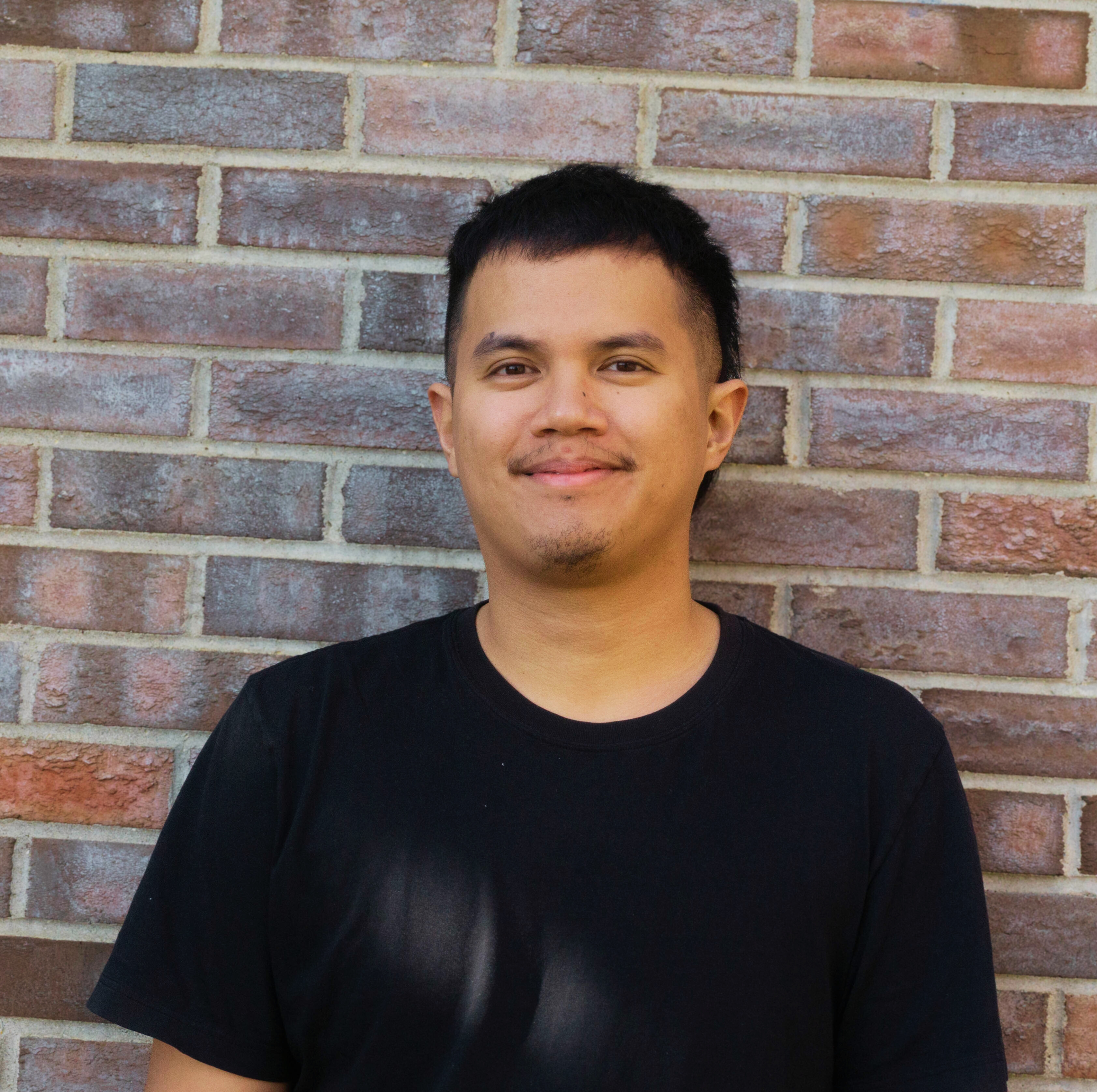 Jose Macasinag, a Filipino person with tan skin and black hair smiling with their mouth closed at the camera. He is wearing a black shirt. The background is a red brick wall.