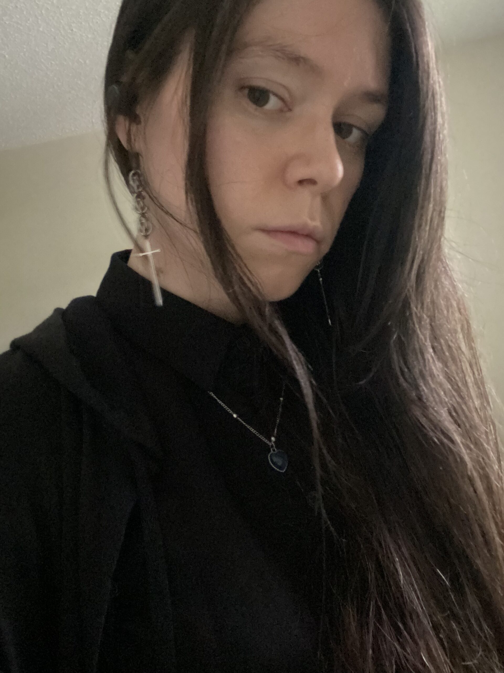 Javiera Díaz, a light skinned person with long dark hair. They are wearing a black blouse and a sword earring in her right ear. They are looking at the camera.