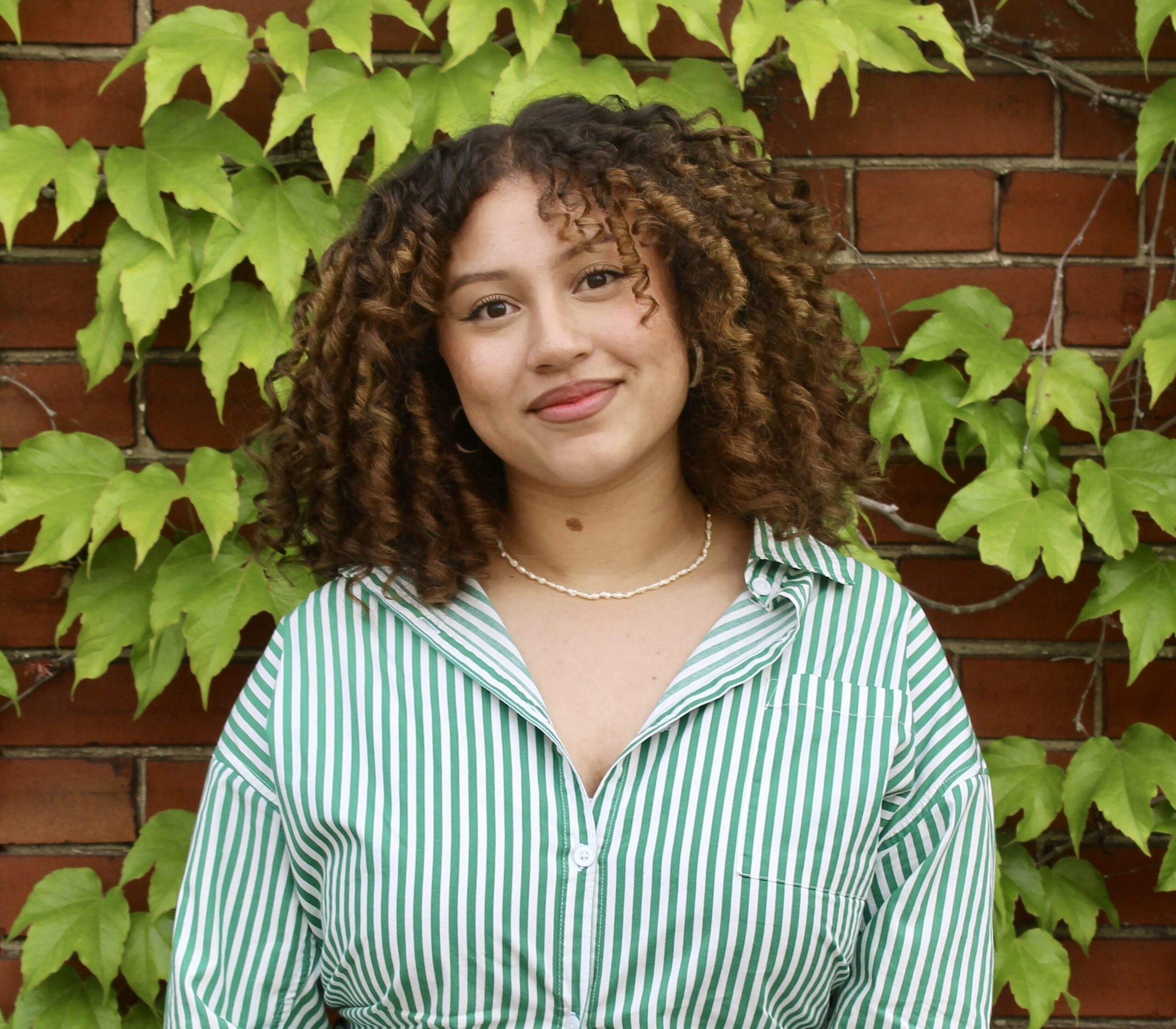 B Roett, a Caribbean Southeast Asian artist stands against a brick wall covered in lush green ivy. They have brown curly, shoulder-length hair with a soft, warm smile directed at the camera. They wear a green and white striped button-up shirt, around their neck is a simple white beaded necklace.