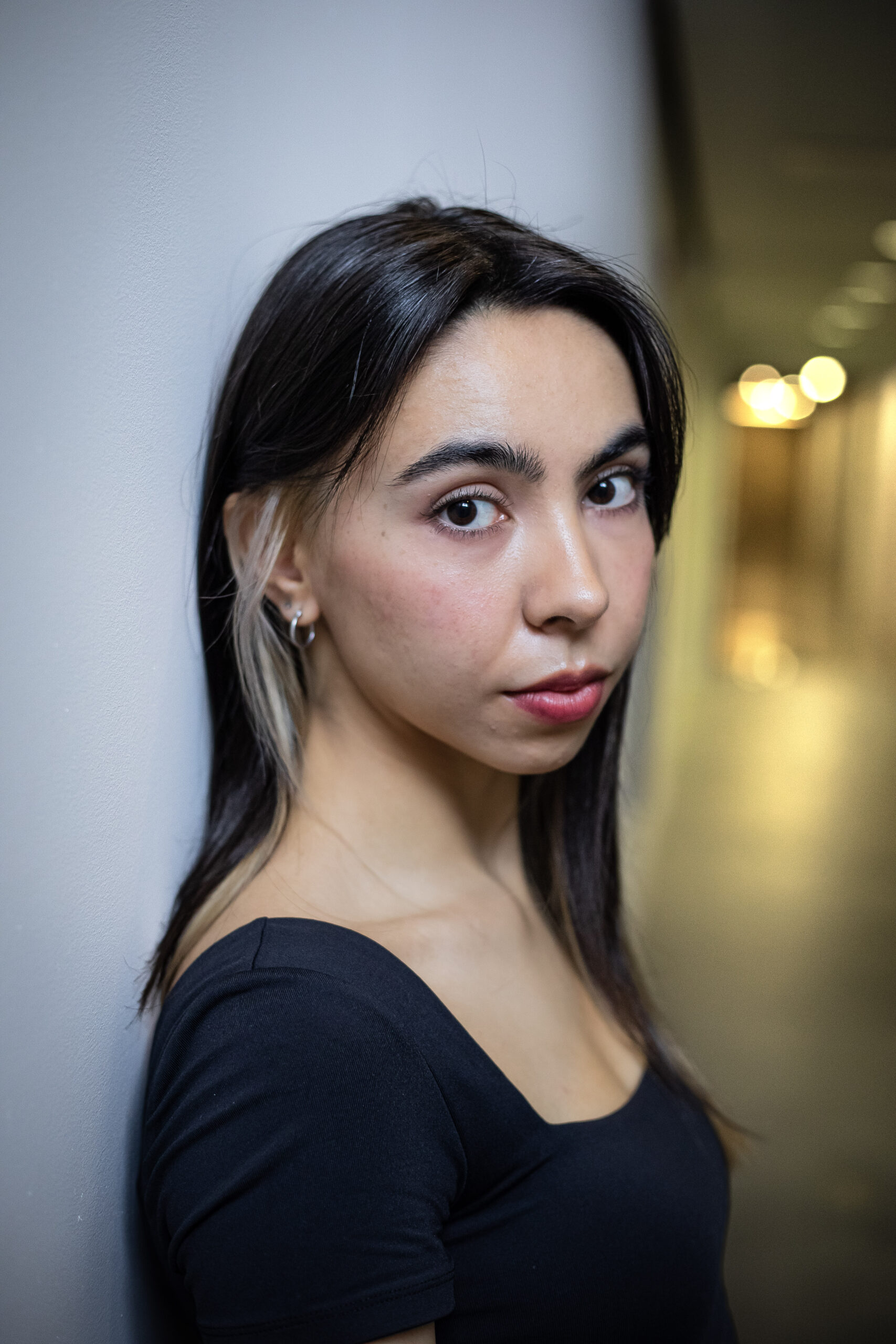 Felicia Holmes, half-Filipino woman with tan skin, brown hair with bleached hair by the ear, looking dramatically at the camera. She wears a black top. The background is white with a yellow lit hallway to her right.