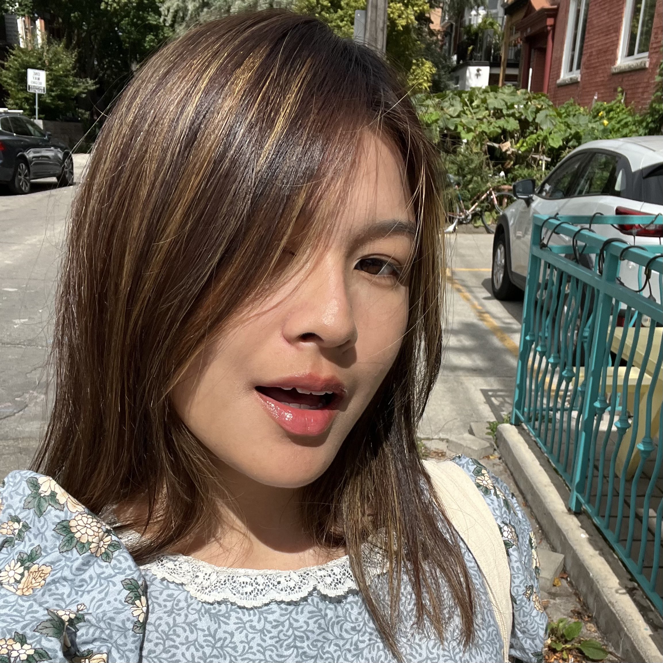 Kayli Chow, a Chinese person with tan skin and dark brown hair smiling at the camera. She is wearing a dusty blue dress with a white flower pattern. The background is a street with foliage.