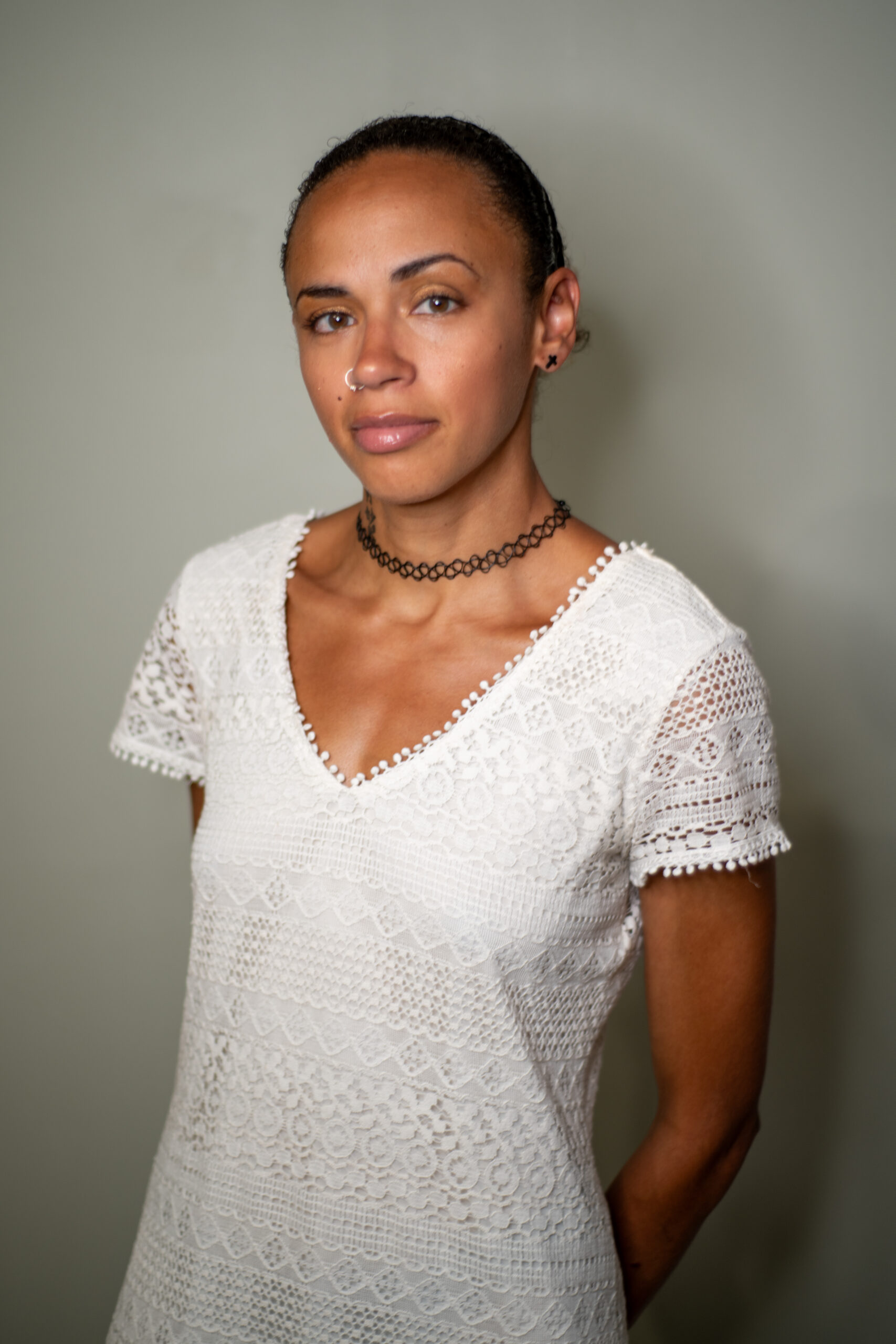 MelVee X, a mixed race Black woman with light brown skin, with her hair pulled back into a bun wearing a white dress with floral texture and a golden M necklace. She is looking directly into the camera, smiling slightly with her left eyebrow raised.