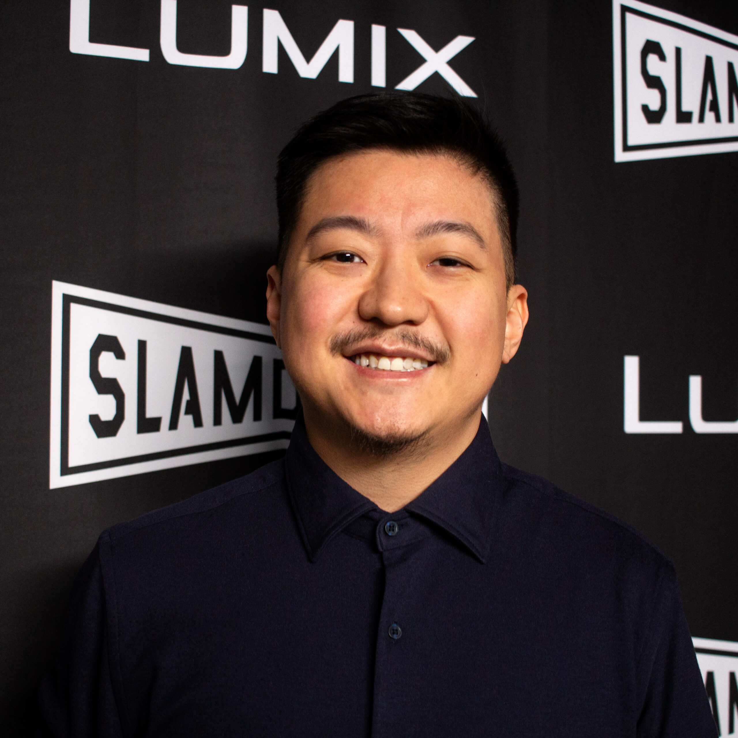 A headshot of Mike Yi Ren, an Asian man with short, neatly styled black hair and a trimmed mustache and goatee. He is smiling warmly at the camera, wearing a dark, buttoned-up collared shirt. The background features a black wall with white logos from the Slamdance film festival.