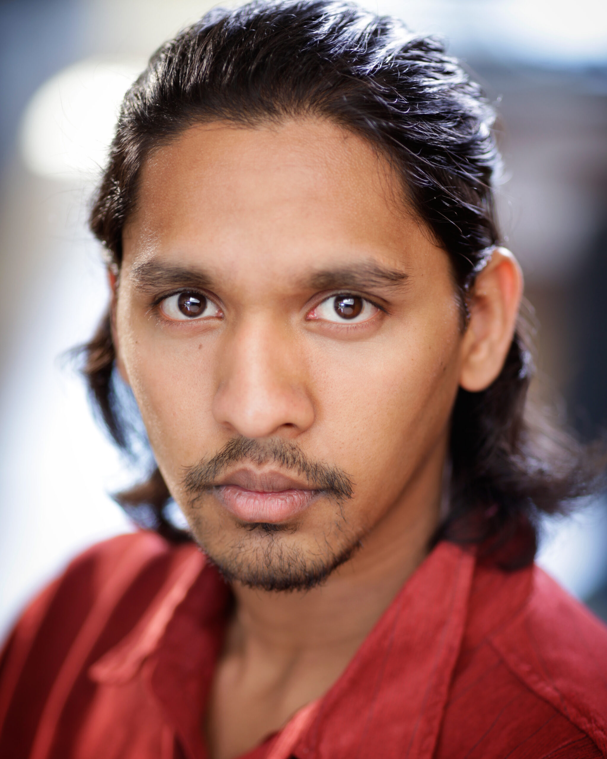 Suchiththa Wickreemsooriya, a multi-ethinic, lighter-brown-skinned Sri Lankan man with tied back black hair, stares neutrally at the camera. He is wearing a red collared shirt. The background is out of focus, but appears to be outdoors.