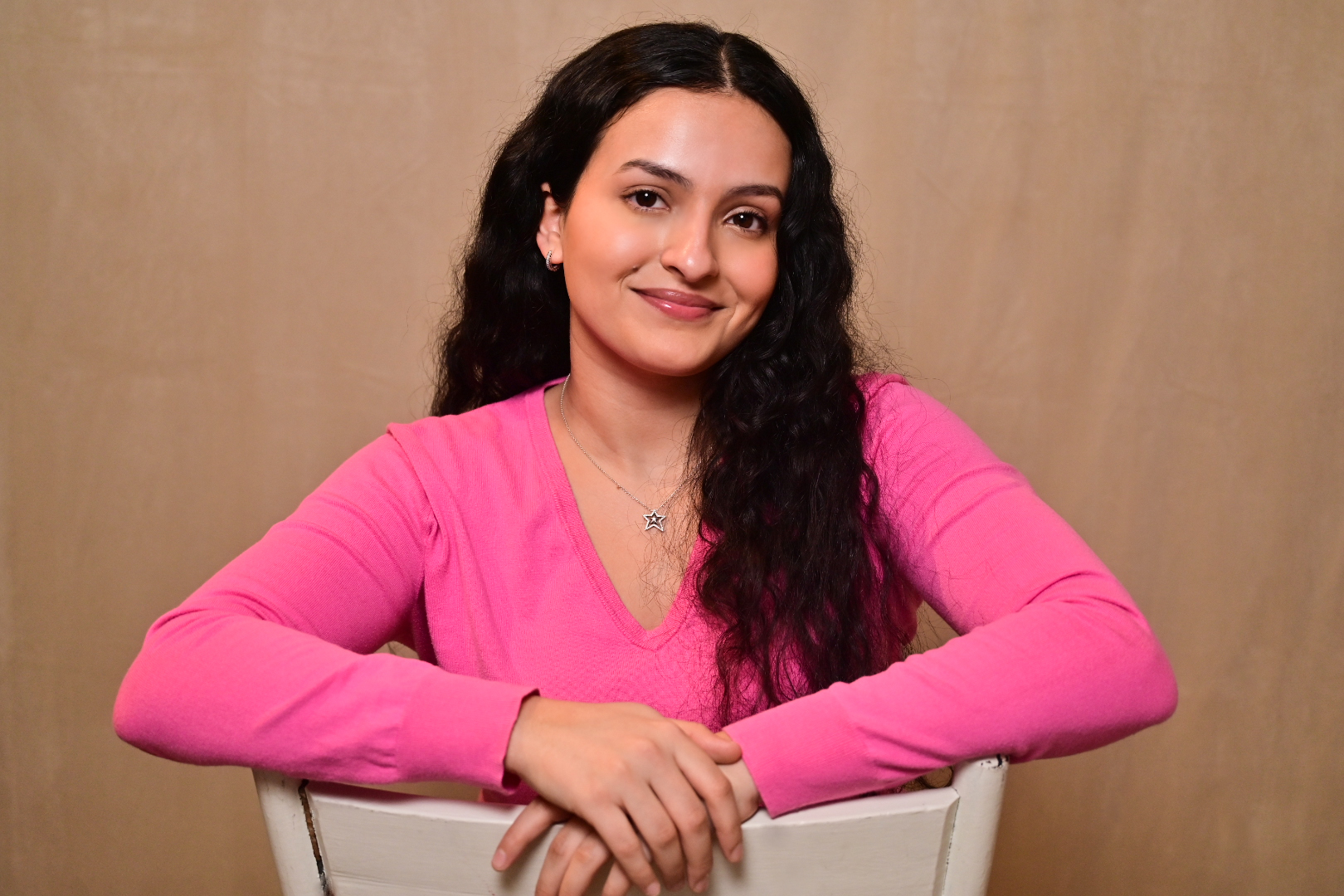 Zoe Marín, a woman with light skin and long, curly black hair smiles to the camera with her mouth closed. She is wearing a pink v-neck and a silver ‘star’ necklace. Her arms are draped over the back of a white chair.