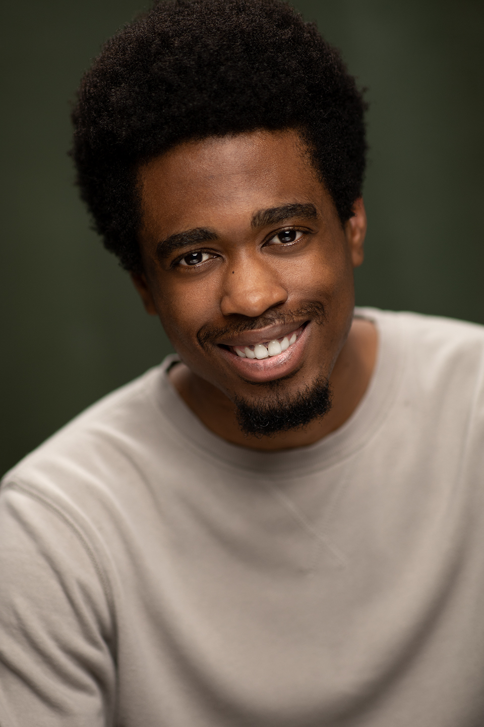 Chisom Akomas, a Nigerian person with brown skin and a mini black Afro, he smiles showing his teeth at the camera. His goatee isn’t fully connected. He’s wearing a beige long sleeve sweater. The background is dark green.