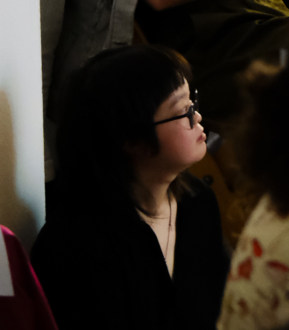 Cyril Chen, a Chinese person with light skin and brown-black hair looks to the right. They are wearing a black v-neck dress with a necklace that has a black thread. They are sitting amongst an audience, watching a screening at Neutral Ground Artist Run Centre. Other audience members are wearing colourful clothes, ranging from mustard to red velvet, and denim.