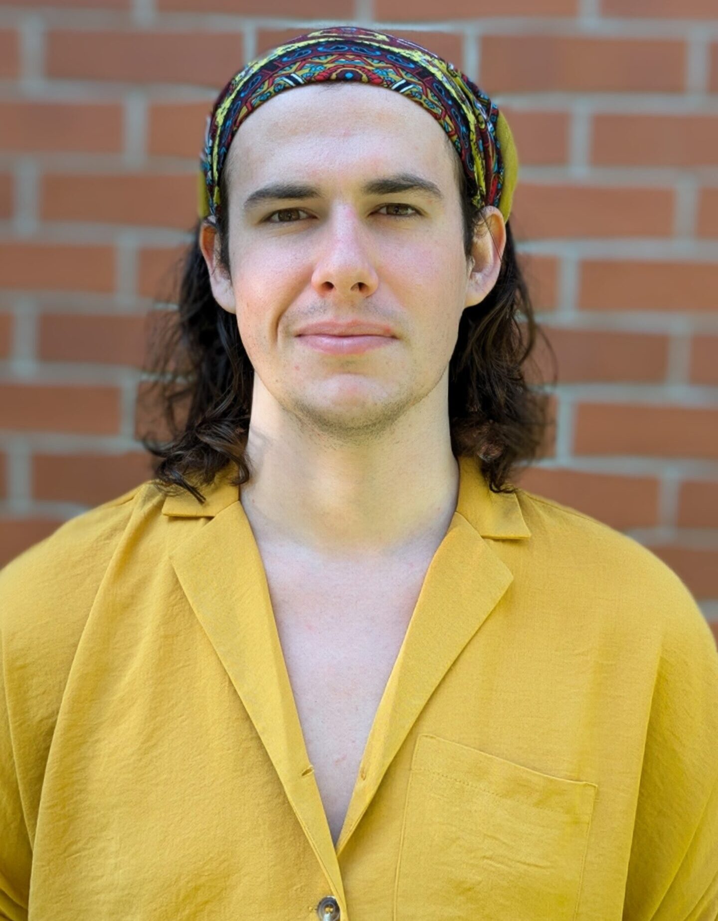 Jacob Loat, a white individual with medium-length brown hair and a yellow, button-up shirt. They are wearing a bandanna.