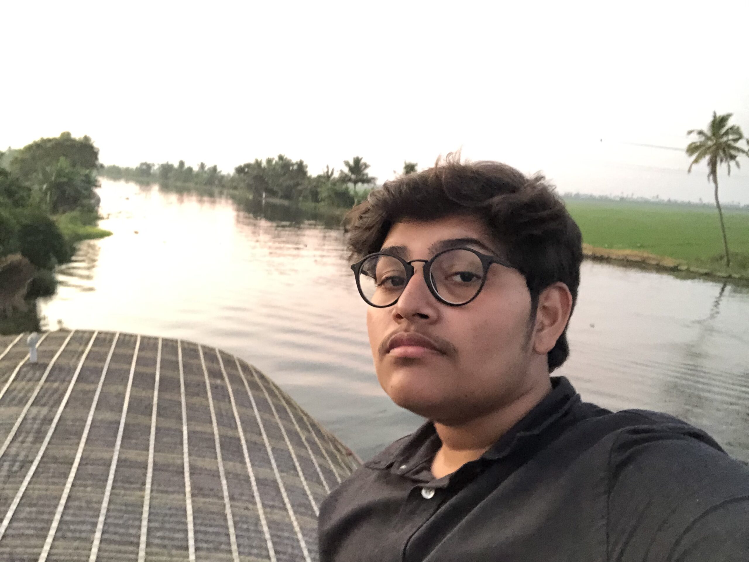 Percy Ali, a Singaporean-Indian person with light brown skin and short, dark hair wears round black glasses and looks at the camera with a calm expression. He is dressed in a dark shirt and are standing on a boat on a river. The background includes lush green fields, palm trees, and a still river, with a soft pinkish hue in the sky.