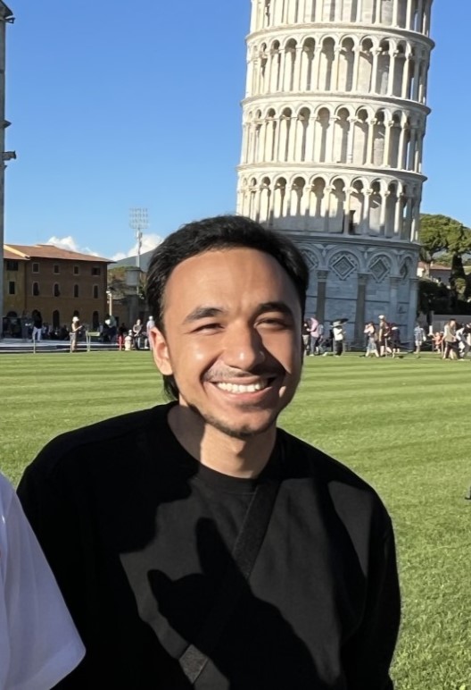 Isaiah Wuthrich, an Asian person with light-tan skin and short black hair smiles at the camera. He is wearing a black sweater with a strap around his shoulder. Behind him is a field and a crowd surrounding a large tower.